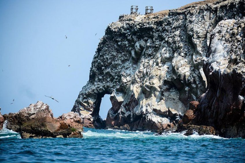 A rocky coastline with a large natural arch formation and a few people standing on the cliff. 4