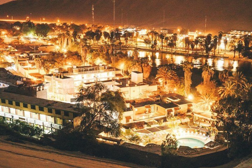 A nighttime view of a city with illuminated buildings, a body of water, and palm trees. 1