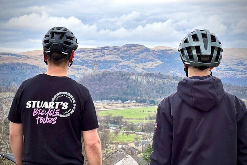 Twee mensen met helmen en fietspakken, met uitzicht op een bergachtig landschap met een verre zicht op een stad. 5