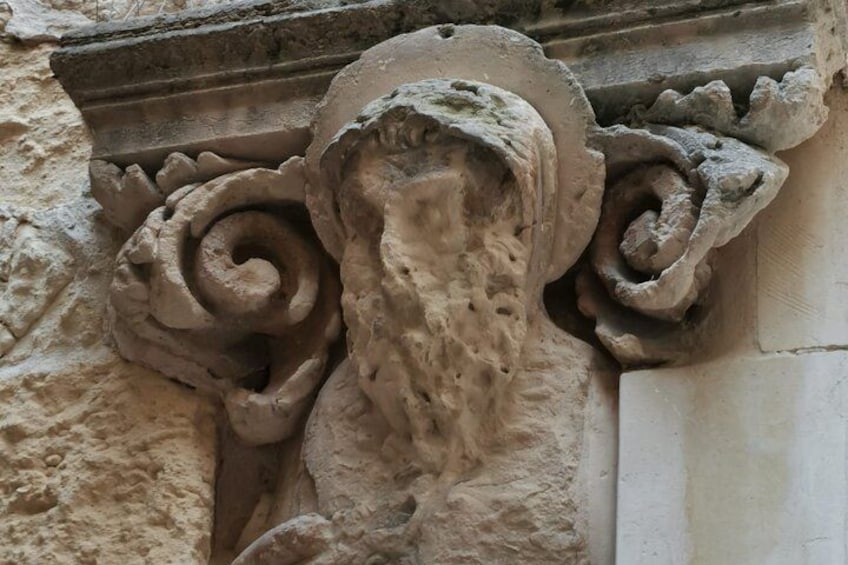 Een stenen beeldhouwwerk van een leeuwenkop met baard, geflankeerd door twee spiraalmotieven, geplaatst in een stenen muur. 5