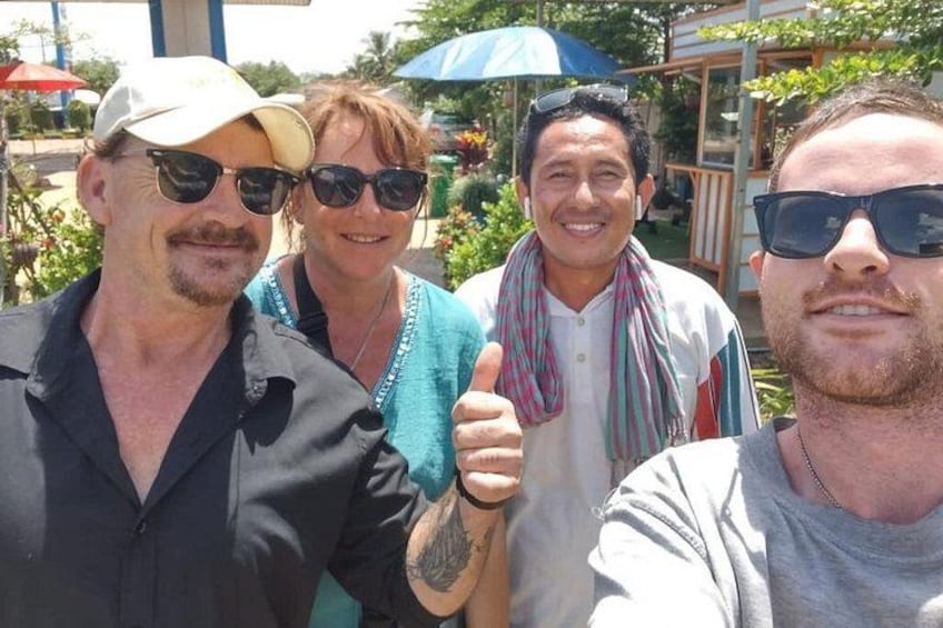 Four people outdoors, one giving a thumbs-up, with a blue umbrella and greenery in the background. 1