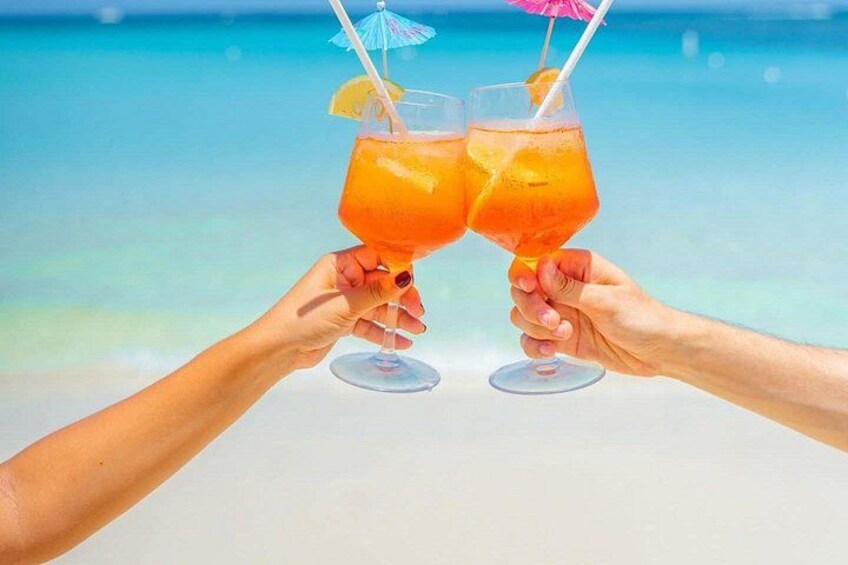 Due mani che sorreggono bicchieri con una bevanda tropicale, con una spiaggia e l'oceano sullo sfondo. 3
