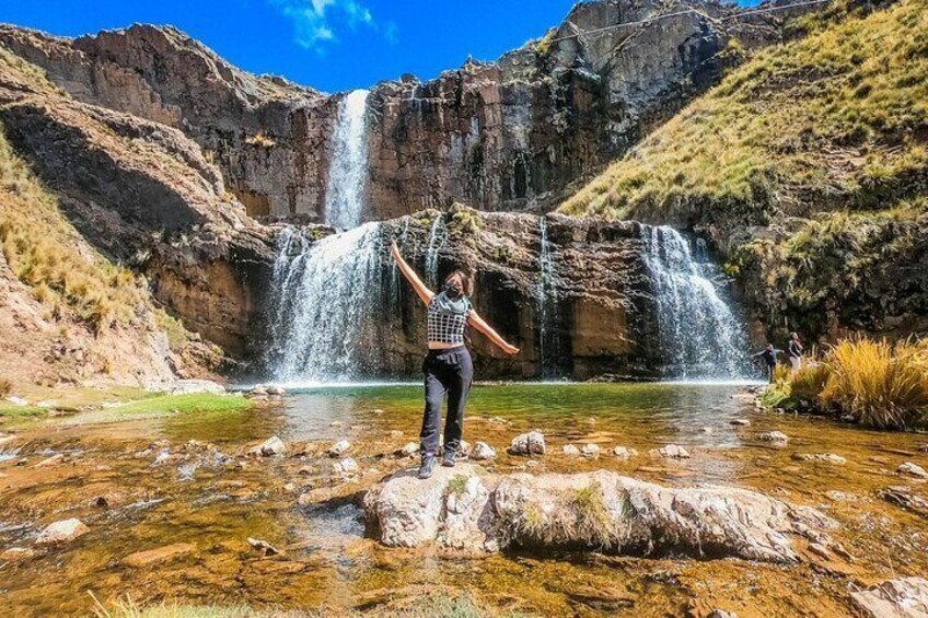 A person standing on rocks in a clear stream with a waterfall in the background. 1
