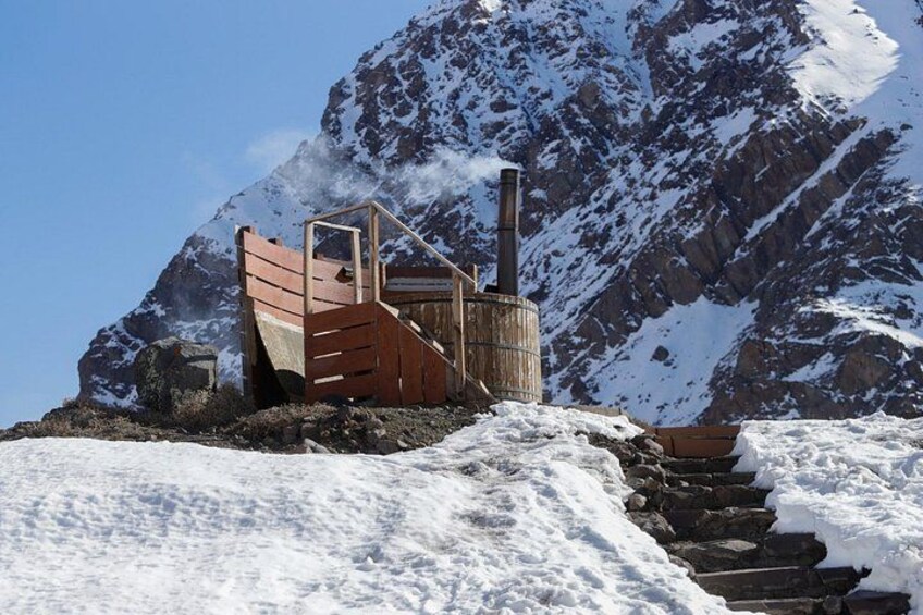 A wooden cabin with a chimney, surrounded by snow-covered mountains and rocky terrain. 3