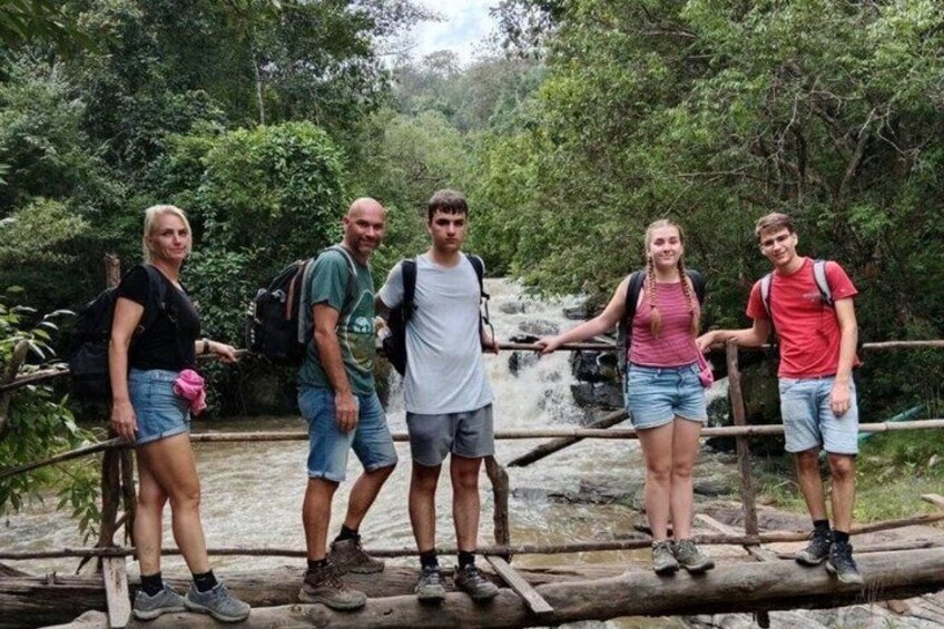 A group of people standing on a wooden bridge over a waterfall in a forested area. 1