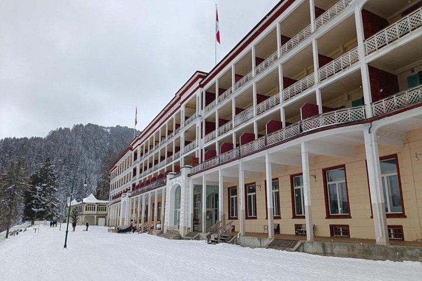 A large, multi-story building with balconies and a flag, surrounded by snow-covered trees and a mountainous backdrop. 3