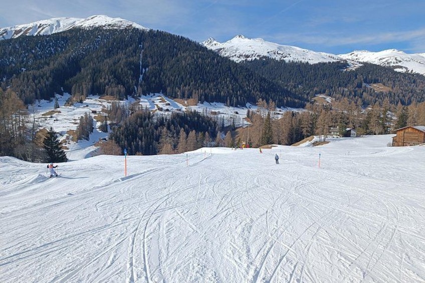 A snow-covered ski slope with skiers, a forested mountain range, and a clear sky. 2
