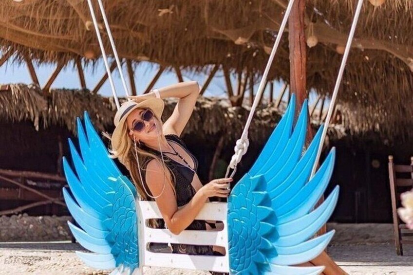 Una persona su un'altalena con ali blu, un ombrello di paglia e uno sfondo con spiaggia. 4