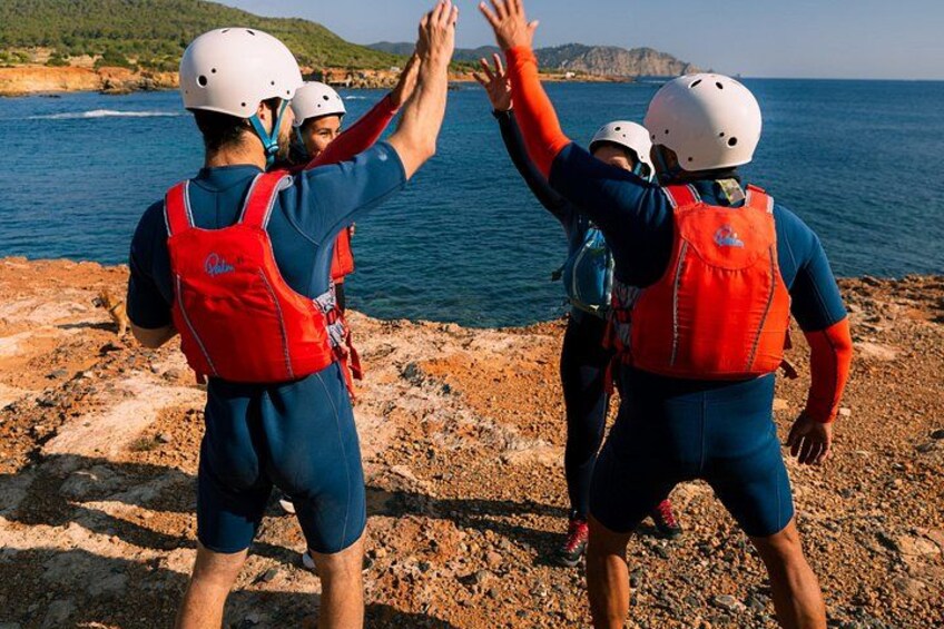 Three people wearing helmets and life jackets, standing on a rocky shore with the sea in the background. 2