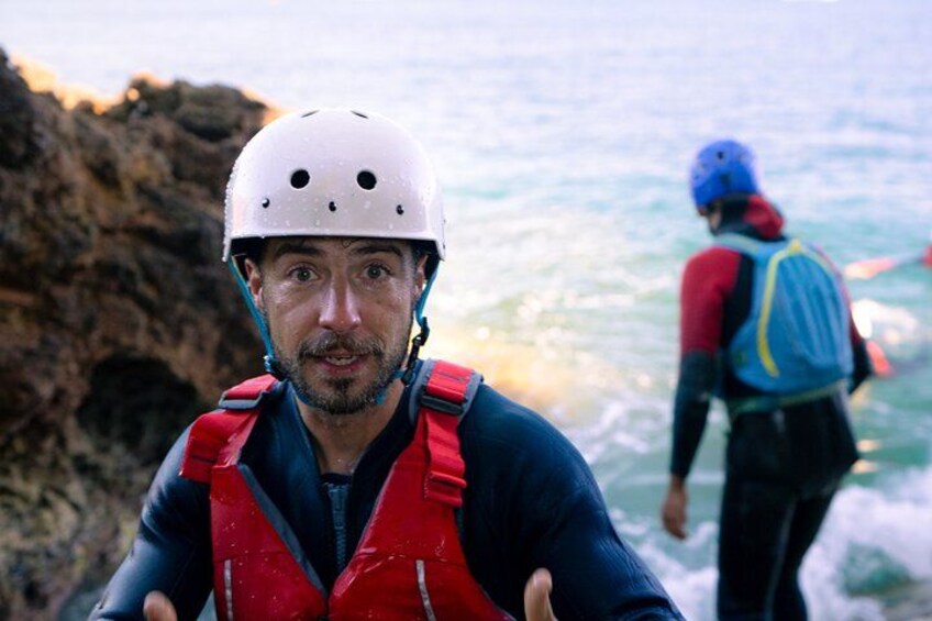 Eine Person mit weißem Helm und roter Schwimmweste in der Nähe von felsigem Gelände, im Hintergrund eine weitere Person. 5