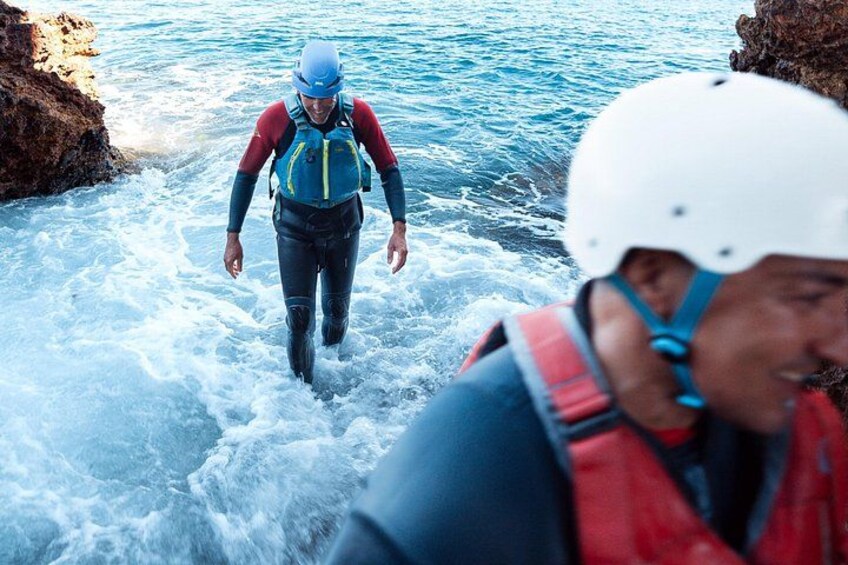 Two individuals in water with helmets and life vests, walking towards rocky shores. 4