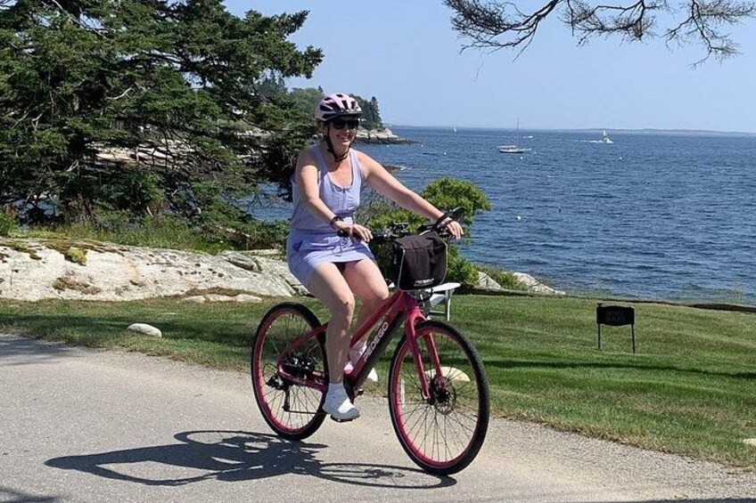 A person riding a bicycle on a paved path near a body of water with boats in the distance. 3