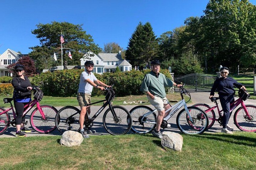 A group of people with bicycles in a park with grass, rocks, and houses in the background. 5