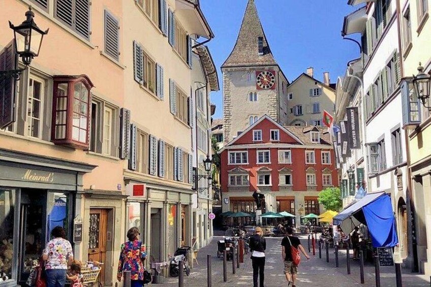 A European street with buildings, a clock tower, and pedestrians. 1