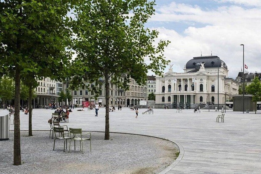 A public square with a large building in the background, trees, and people walking. 3