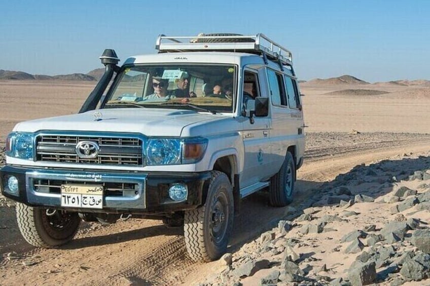A white Toyota Land Cruiser with off-road tires, a roof rack, and a license plate in a desert landscape. 5