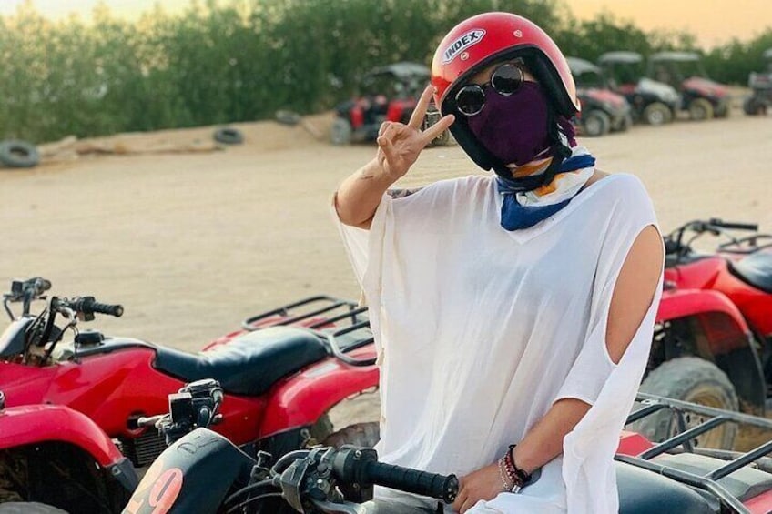 A person wearing a white shirt and a red helmet, standing next to a red ATV in an open area with other ATVs in the background. 1