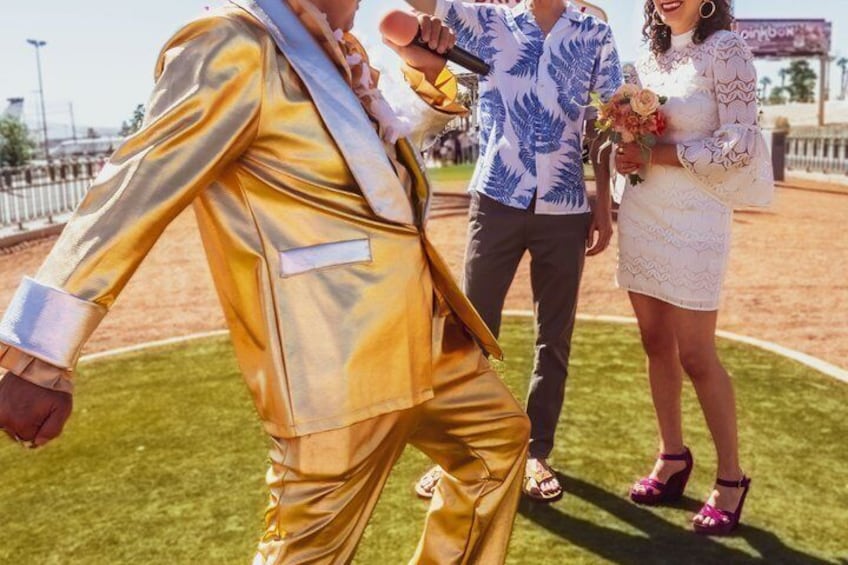 Eine Person in einem Goldsanzug, eine weitere in einem blau gemusterten Hemd und eine Frau in einem weißen Kleid, die einen Blumenstrauß auf einer Wiese hält. 5