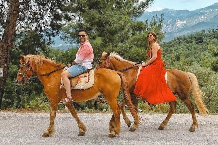 Dos personas montando a caballo por un camino pavimentado, con árboles y montañas al fondo. 1