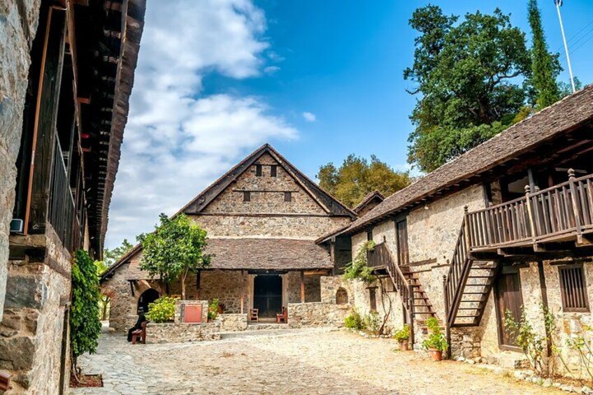 Une cour pavée de pierres, bordée de bâtiments en pierre aux allures rustiques, de balcons en bois et surplombée d’un ciel d’un bleu éclatant. 1