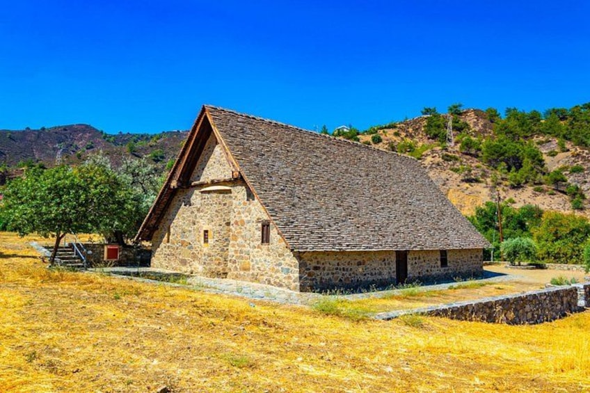 A stone building with a sloped roof in a dry, grassy field with trees and hills in the background. 2