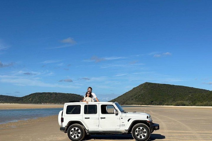 En hvid SUV parkeret på en sandstrand med en person siddende på motorhjelmen, en klar blå himmel og en bakke i baggrunden. 5