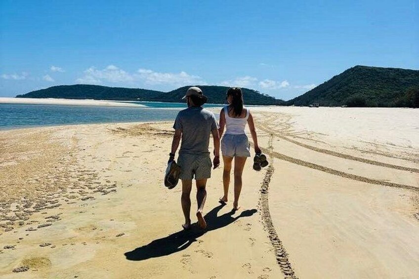 Two people walking on a sandy beach with footprints, a clear sky, and distant hills. 4