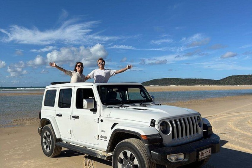 En hvid Jeep på en strand med to personer stående på taget, med armene strakt ud. 4