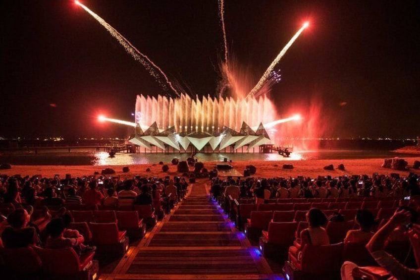 A nighttime outdoor event with a large audience seated in rows facing a stage with water fountains and lighting effects. 5