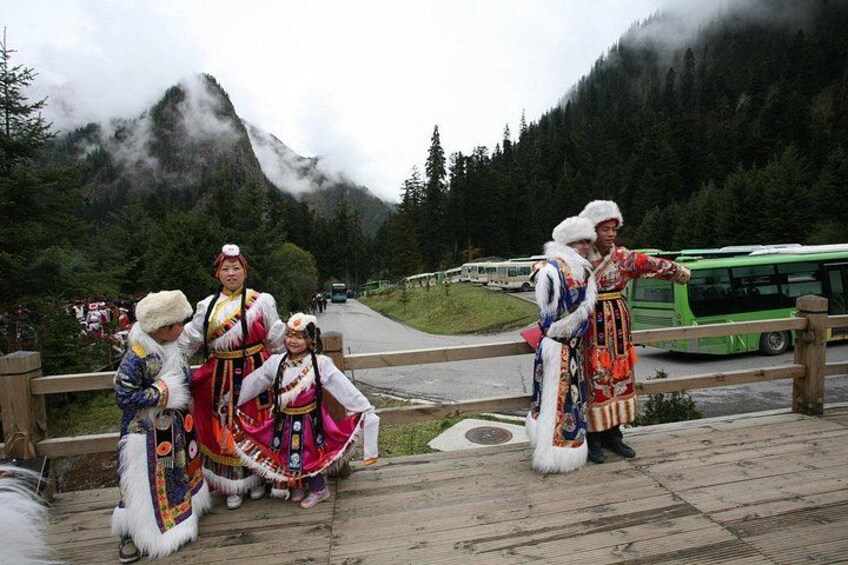 Un groupe de personnes, vêtues de costumes traditionnels, se tient sur une terrasse en bois. En arrière-plan, on aperçoit un paysage montagneux et un autobus vert. 4