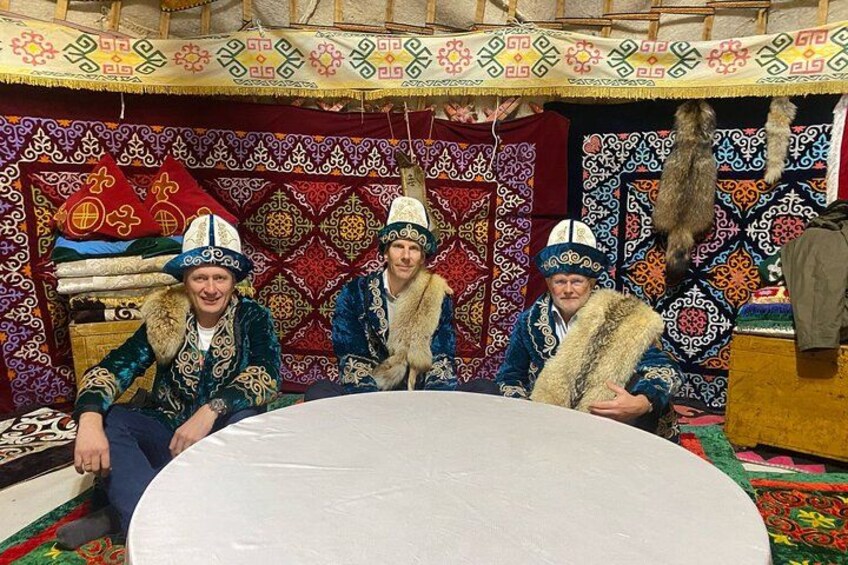 Three individuals seated at a round table in a tent with patterned fabric walls and a fur-lined bench. 1