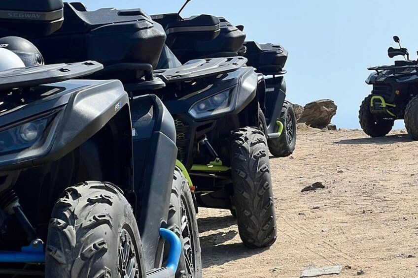 A row of ATVs parked on a dirt surface with a rocky outcrop in the background. 2