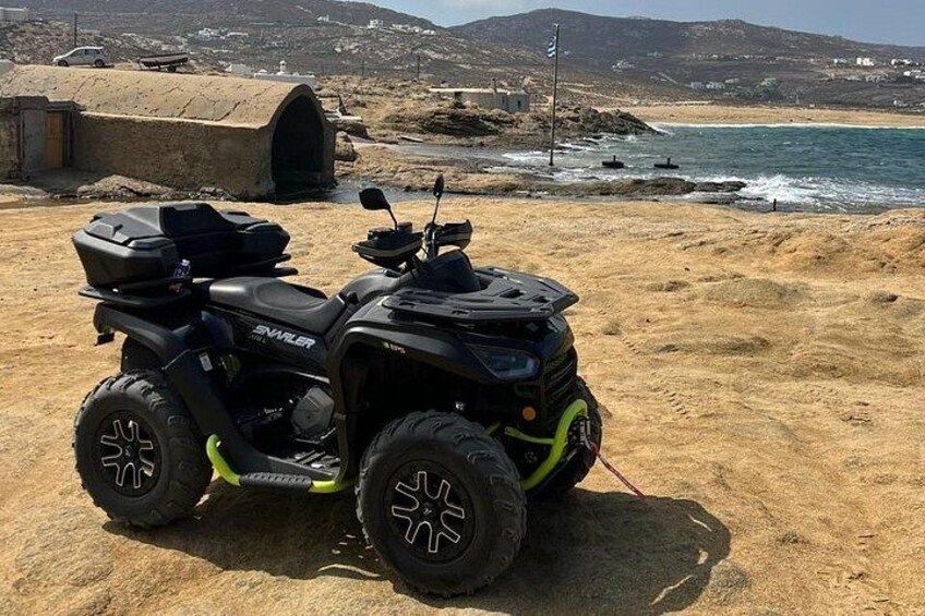 A black ATV with a green light on the front wheel, parked on a sandy beach with a rocky outcrop and the ocean in the background. 2