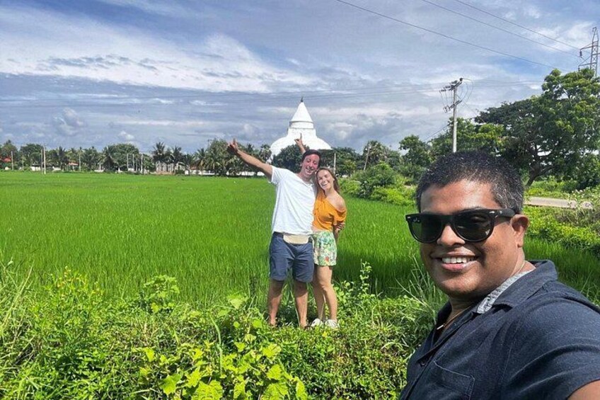 A person taking a selfie with a white structure in the background, a green field, and a clear sky. 2