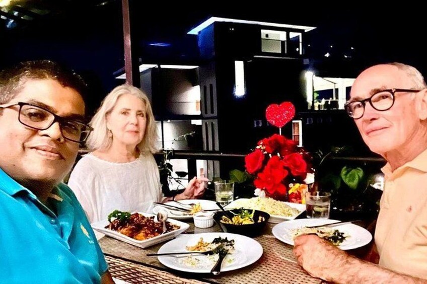 A group of people dining outdoors at night with a table set with plates of food and a vase of red flowers. 3
