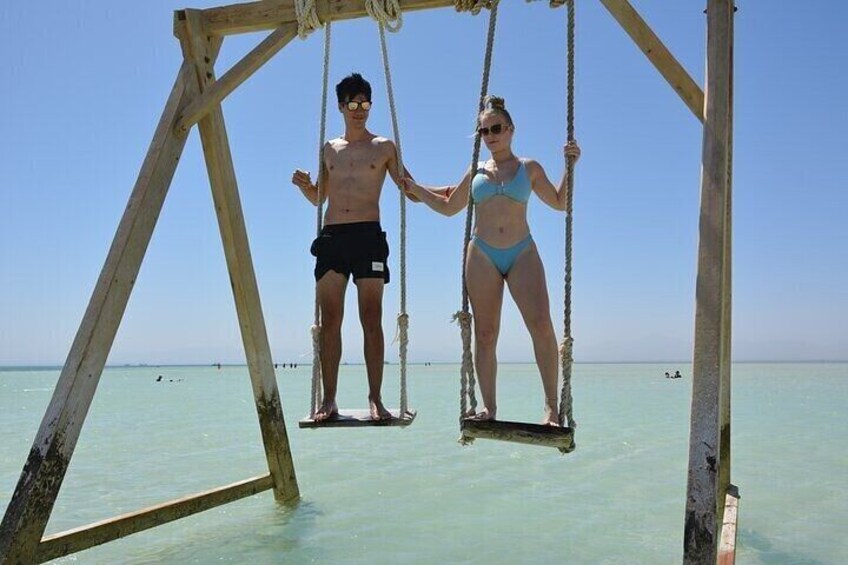 Twee personen op een houten schommel boven helderblauw water. 3