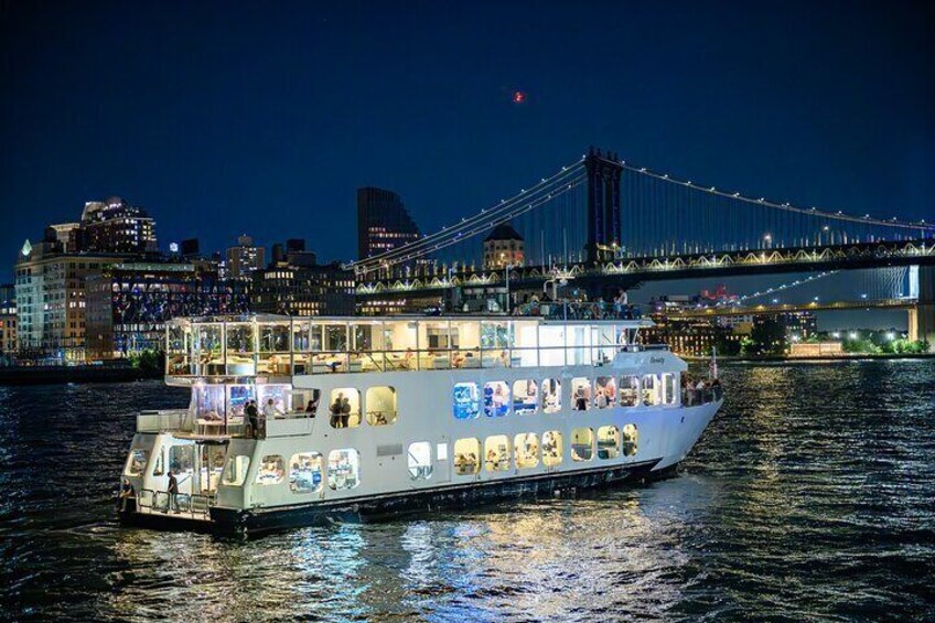Un bateau de croisière fluviale, doté de plusieurs ponts et transportant de nombreux passagers, est amarré près de la silhouette d’une ville, avec un pont imposant en arrière-plan. 1