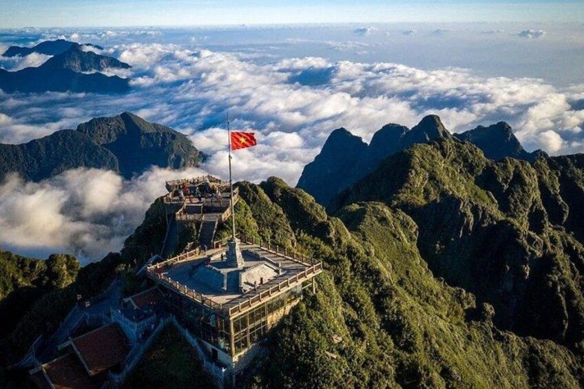 Un pico montañoso con una bandera, vegetación exuberante y un cielo despejado. 1