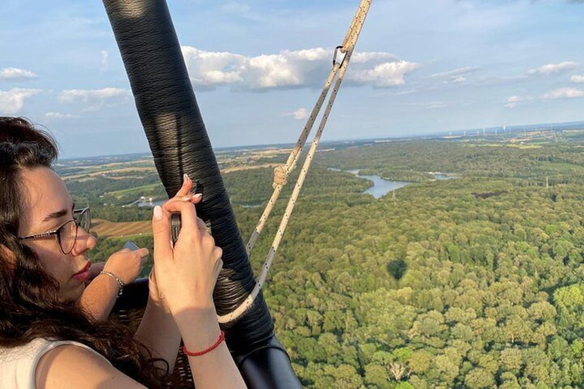 A person holding onto a cable, overlooking a forested landscape with a river and distant wind turbines. 2