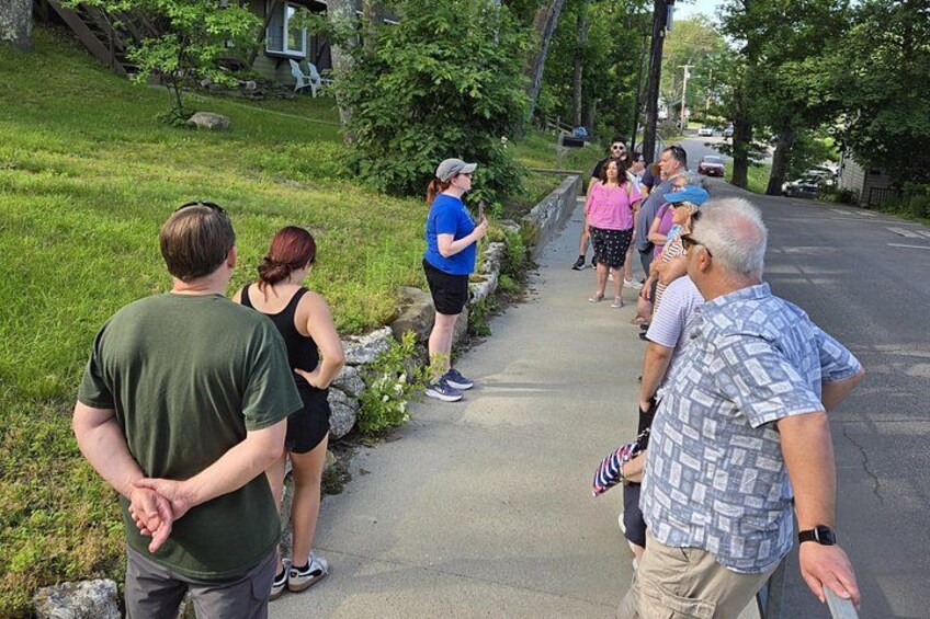 A group of people standing on a sidewalk in a residential area with houses and trees in the background. 3