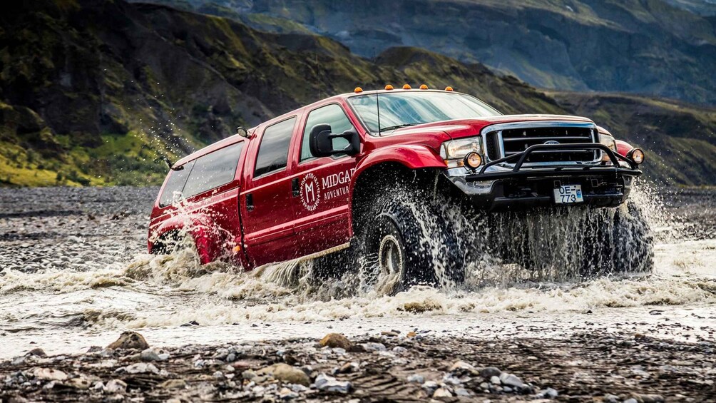 A red truck driving through a shallow river, surrounded by rocky terrain and mountains. 2