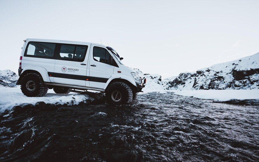 A white van with 'MIDGARD ADVENTURE' written on it, driving through a snowy landscape with a river. 4