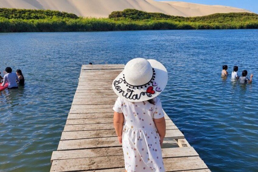 A wooden dock extending into a calm body of water with people swimming and standing in the background. 4