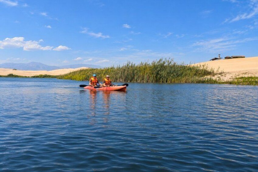Two people kayaking on a calm river with sand dunes and vegetation in the background. 2