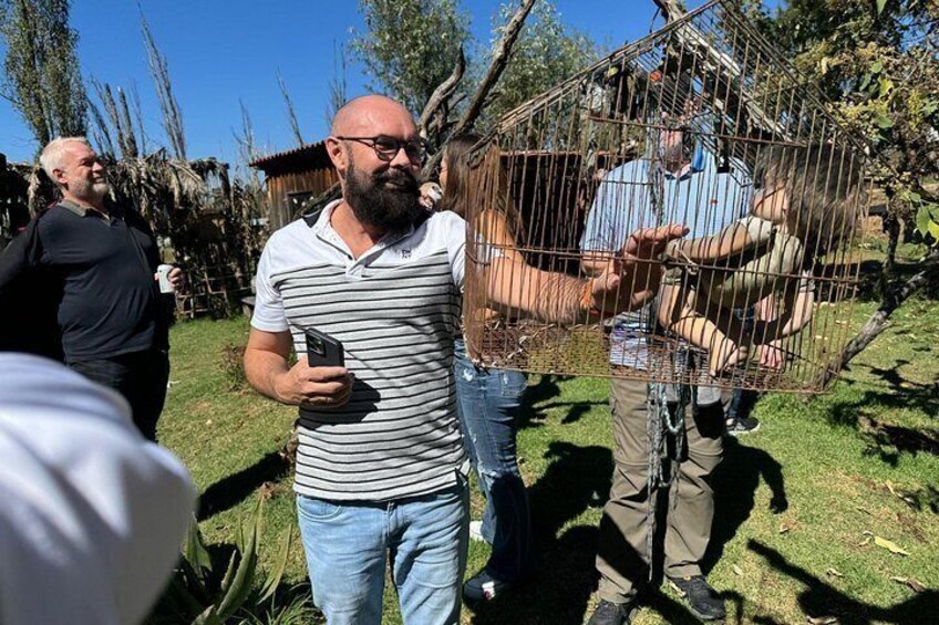 Un homme vêtu d’un t-shirt rayé et d’un jean, debout à l’extérieur, tenant un téléphone. On aperçoit, à l’arrière-plan, une cage en métal et une personne à l’intérieur. 3