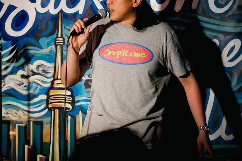 A person holding a microphone, wearing a grey t-shirt with a logo, standing in front of a colorful mural with a tower-like structure. 3