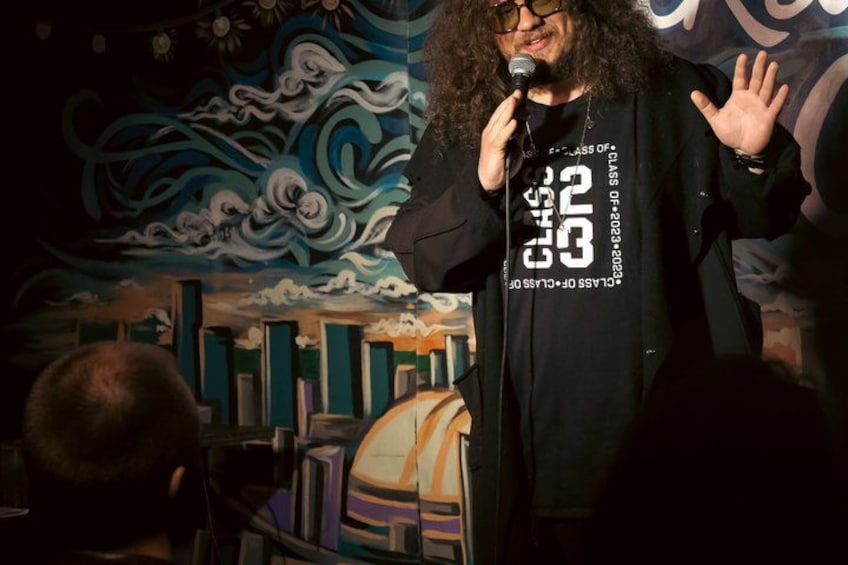 A person with curly hair, wearing a black t-shirt with white text, speaking into a microphone. 5