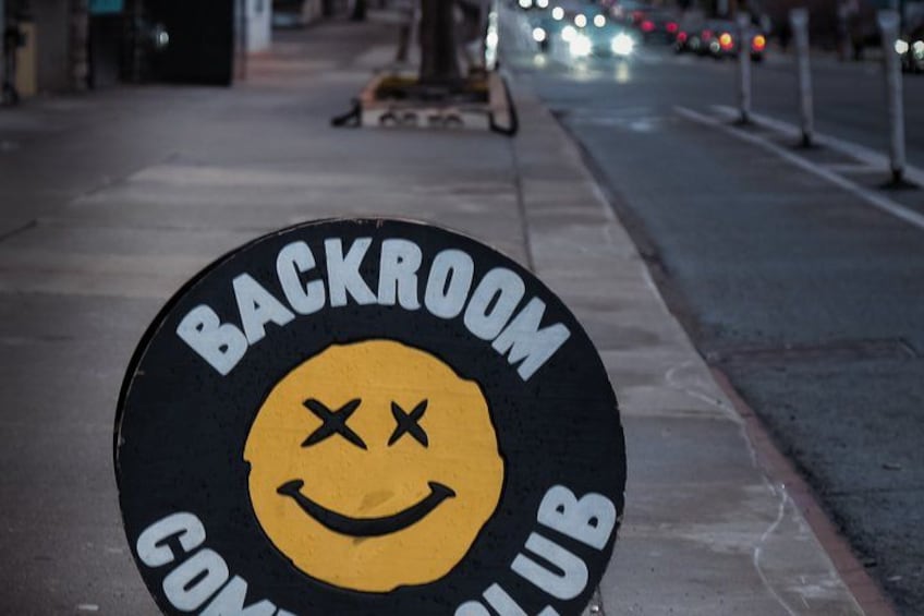 A circular sign with a yellow smiley face and the words 'BACKROOM CLUB' on a sidewalk. 2