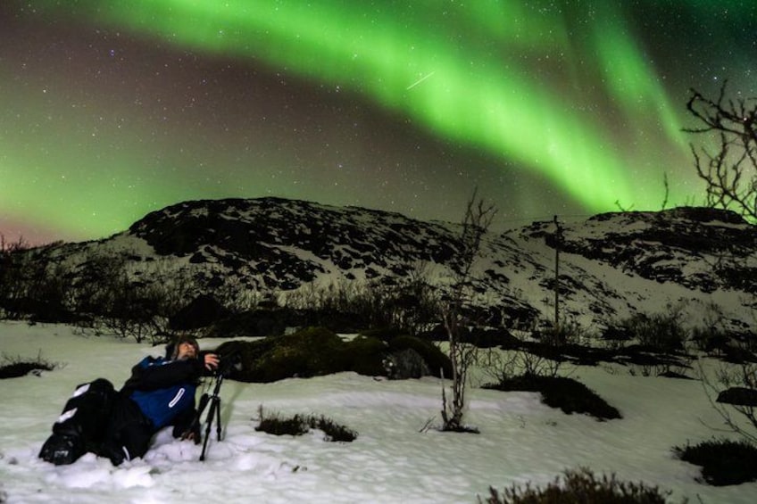 En person ligger i sneen med en cykel under en nattehimmel med grønne nordlys. 2