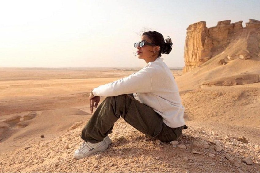A person sitting on a rocky outcrop in a desert landscape with a large rock formation in the background. 1